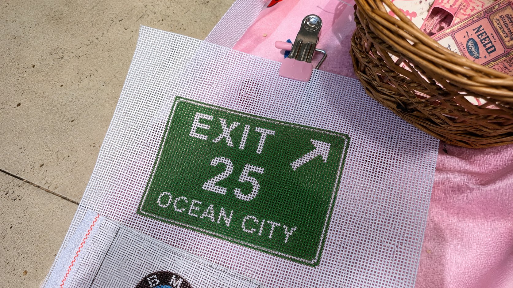 An Ocean City Exit 25 needlepoint canvas on a table beside a basket of canvases and a project bag